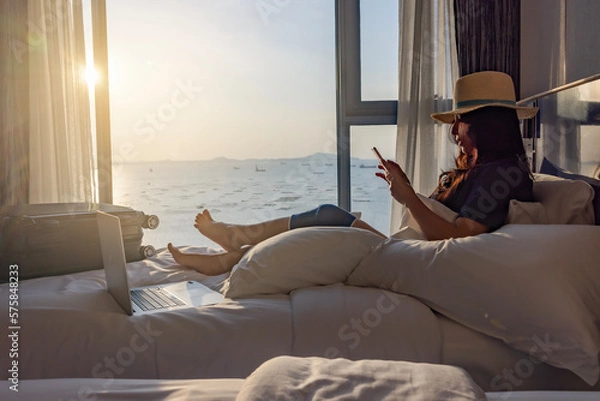 Obraz Travel insurance covers accident and medical expenses. Woman traveler with knee wrapped in medical bandage after travel accident sitting on bed in room, using smartphone and laptop to insurance online