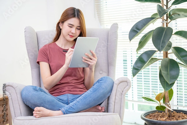 Obraz Asian woman sitting on yellow sofa using digital tablet and pen for learning shopping or working online near window
