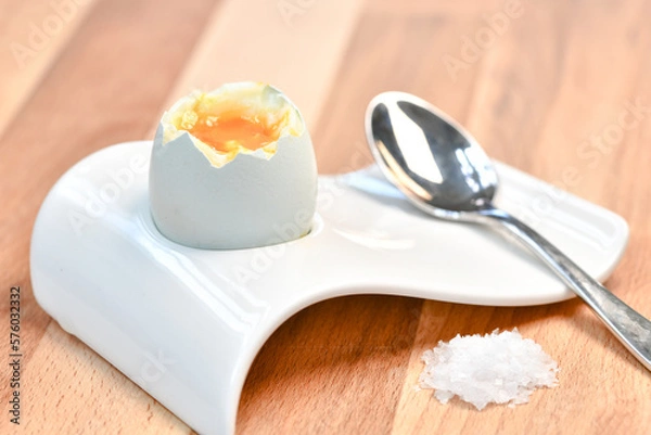 Obraz a single boiled egg in an egg cup broken open showing yolk with a spoon on a wooden surface