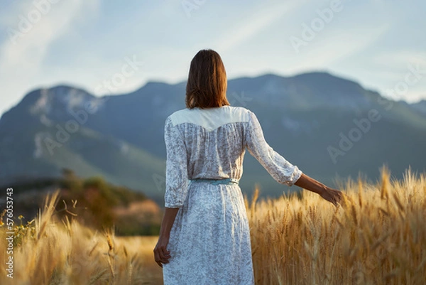 Obraz Rear view elegant woman enjoying nature view of mountains. Lady wearing white dress standing in golden field of barley or wheat crops at sunset