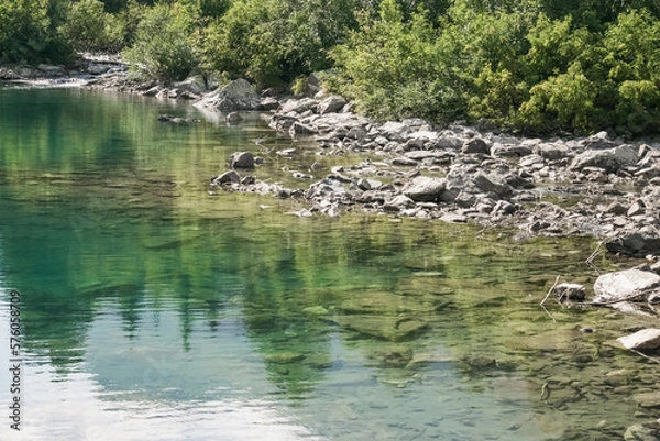 Fototapeta Reflection of treetops and mountain ranges in a turquoise-colored mountain lake with a rocky shore, a landscape on a sunny summer day in the mountains