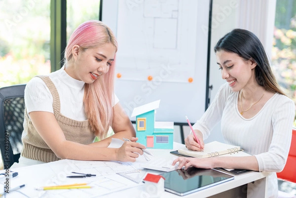 Fototapeta Concept of engineering consulting, Two female engineers discussing about model of building together; Two female architects are studying blueprint of building house.