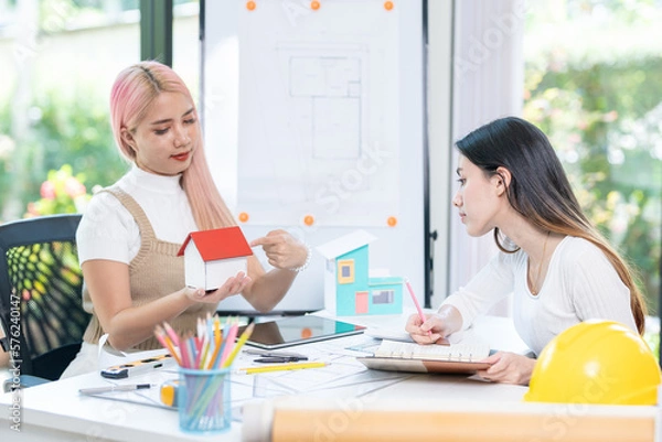 Fototapeta Concept of engineering consulting, Two female engineers discussing about model of building together; Two female architects are studying blueprint of building house.