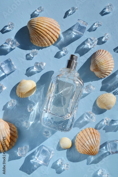 Fototapeta Glass bottle of perfume with seashells, ice cubes, water drops on the light blue background under sun rays. Smell of summer. Fresh  cooling perfumery. Ocean fresh smell. 