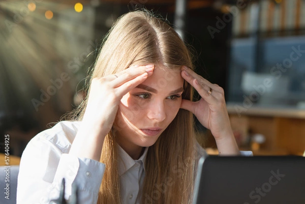 Obraz The girl looks thoughtfully at the laptop monitor. Online work. Freelancer works in a cafe.