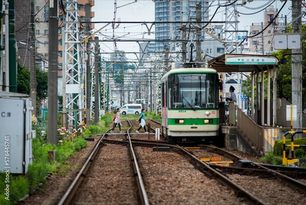 Fototapeta Tokyo Shitamachi trolly train (Toden) running in middle of city and two young asian girls walking across the tracks
