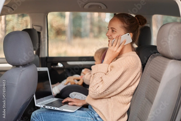 Fototapeta Image of smiling delighted Caucasian woman wearing beige sweater sitting on backseat with her baby and talking smart phone, holding pc computer on her knees.
