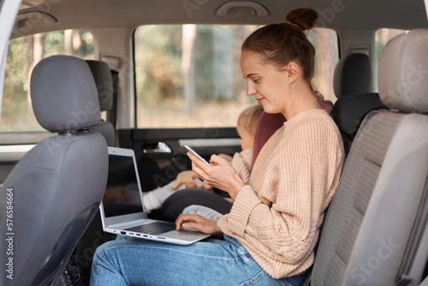 Fototapeta Image of cheerful young woman working on laptop while sitting with her baby daughter in safety chair on backseat of car. female freelancer using cell phone, smiling while looking at phone display.