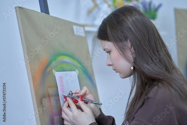 Obraz A teenage girl, an aspiring artist, paints with an airbrush on paper. Drawing lesson in an art studio.