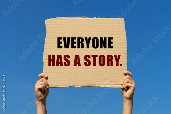 Fototapeta Everyone has a story text on box paper held by 2 hands with isolated blue sky background. This message board can be used as business concept to inform anyone "Everyone has a story.".