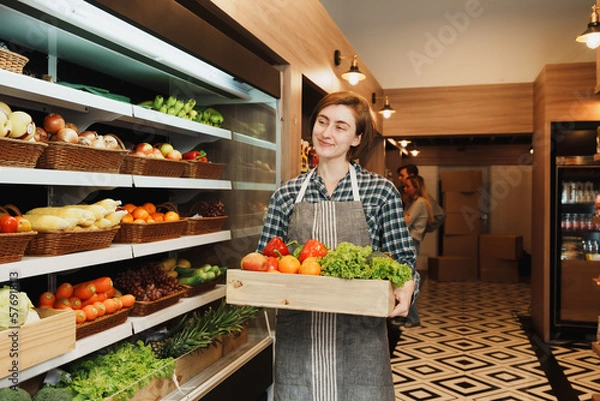 Obraz Caucasian young adult saleswoman is carrying a fruit basket and preparing to add stock of fruit to the shelf. Female employee who working in the grocery store looking at the camera with smile face. 
