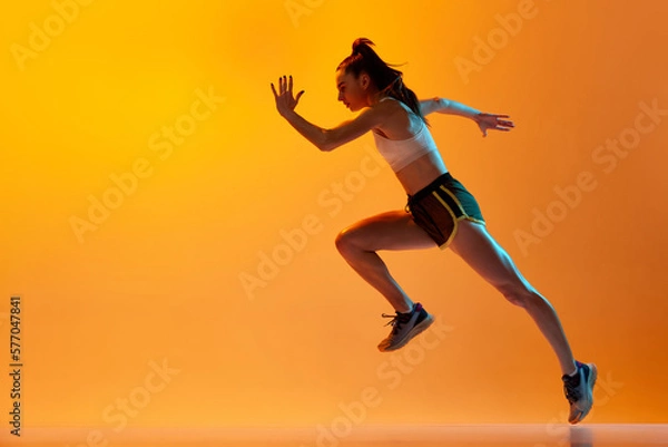 Fototapeta Dynamic image of professional female runner, athlete in motion, running over orange studio background in neon light. Concept of sportive lifestyle, health, competition, endurance, action. Ad