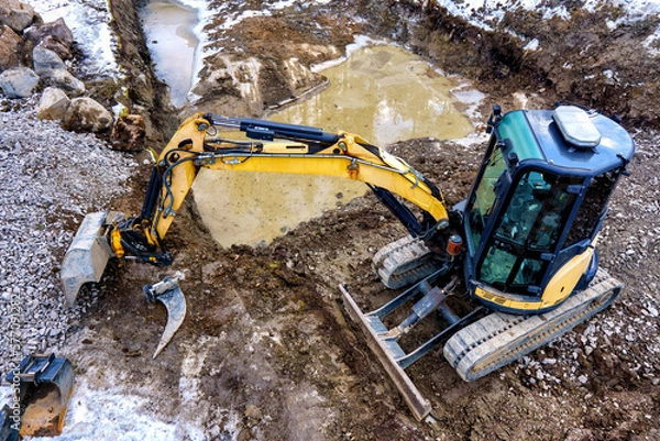 Fototapeta A small yellow excavator on a construction site. Laying of foundations and drainage.
