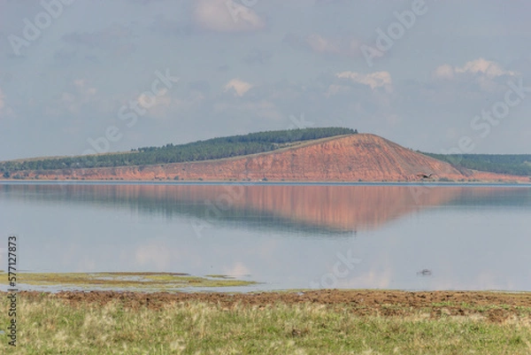 Fototapeta lake with a red hill with forest in the background.gray heron flies and is reflected in the lake