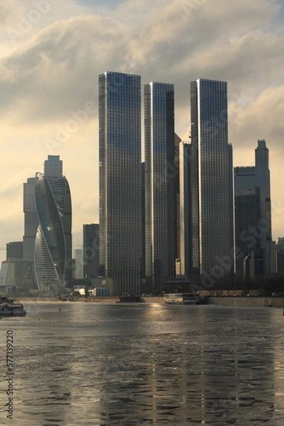 Fototapeta City landscape with a view of the Moscow City skyscrapers and a river with cracked ice in a frosty haze and sun rays on a frosty day.