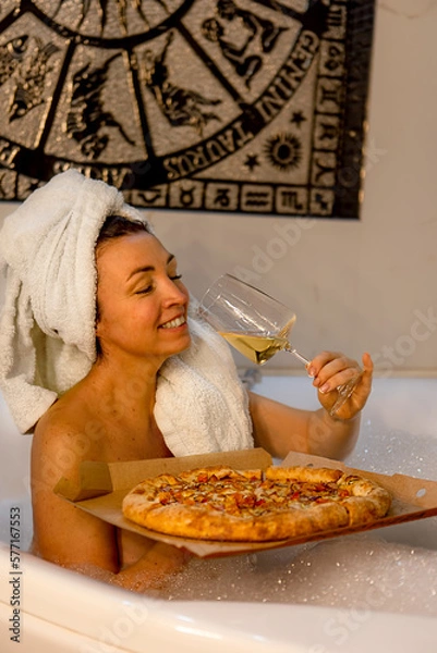 Fototapeta portrait of an attractive dark-haired girl with a towel on her head .the girl sits in the bath drinks wine and eats pizza