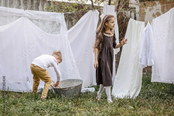 Fototapeta A girl and a boy of primary school age hang their underwear on ropes in the spring in the garden of a country house. the concept of village life, healthy lifestyle, helping mom