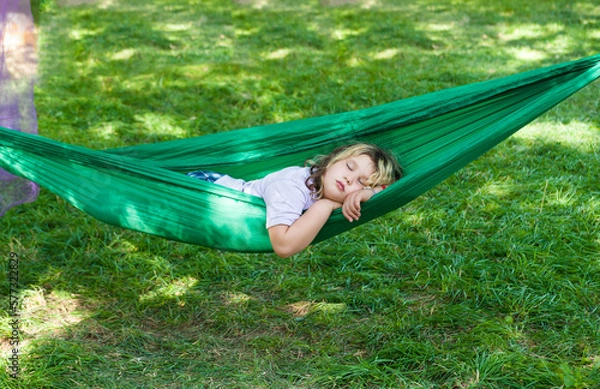Fototapeta A little girl is sleeping in green hammock outside in the forest with bright grass. Summer time nap