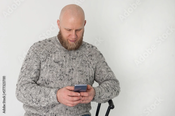 Fototapeta Bold bearded man is sitting on black chair and watching smartphone  in white studio