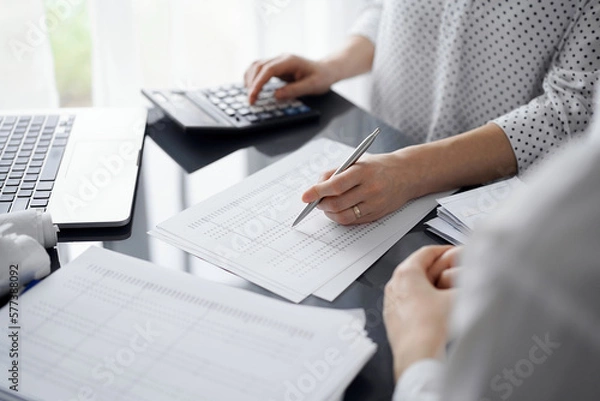 Fototapeta Woman accountant using a calculator and laptop computer while counting taxes for a client. Business audit and finance concepts.