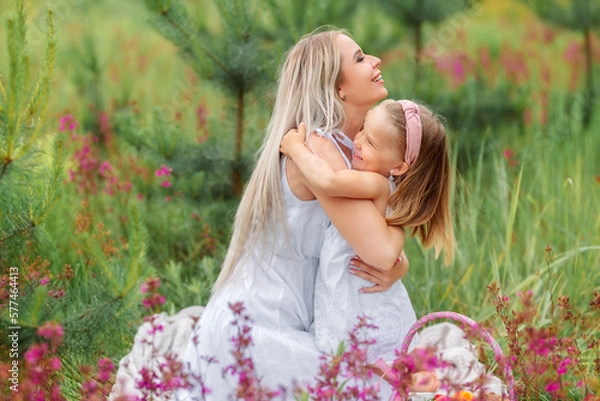 Obraz Mom and daughter on a picnic in nature in summer or spring. They are dressed in white clothes, with flower wreaths on their heads. Around them is a field with grass and flowers.
