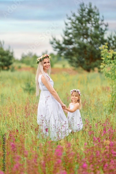 Obraz Mom and daughter in white clothes and wreaths of flowers on their heads walk in a beautiful flower field in summer or spring