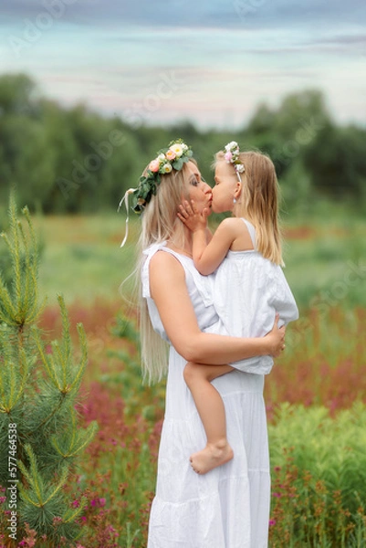 Obraz Mom and daughter in white clothes and wreaths of flowers on their heads walk in a beautiful flower field in summer or spring