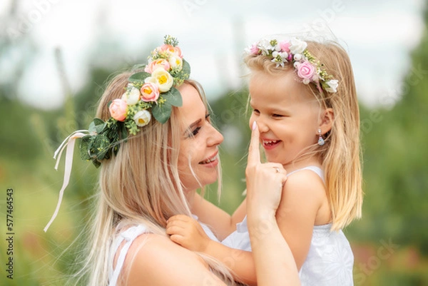Obraz Mom and daughter in white clothes and wreaths of flowers on their heads walk in a beautiful flower field in summer or spring