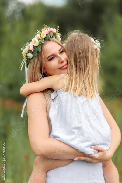 Obraz Mom and daughter in white clothes and wreaths of flowers on their heads walk in a beautiful flower field in summer or spring.