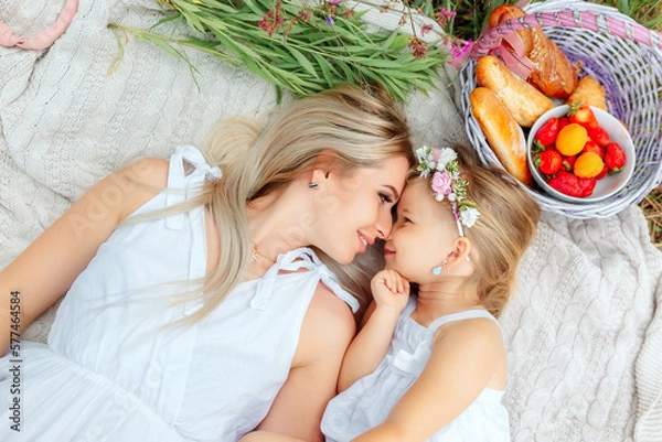 Obraz Mom and daughter on a picnic in nature in summer or spring. They are dressed in white clothes. 
Next to them is a fruit basket and pastries.
