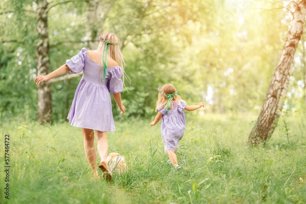 Obraz mother and daughter in short lilac dresses run along a summer meadow with green grass and trees. A small red dog runs in front of them.