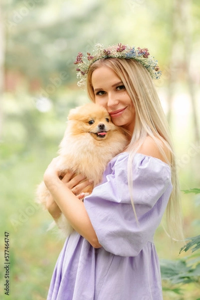 Obraz portrait of a beautiful girl with blond long hair, dressed in a lilac dress and a wreath of flowers. In the hands of a girl a small dog