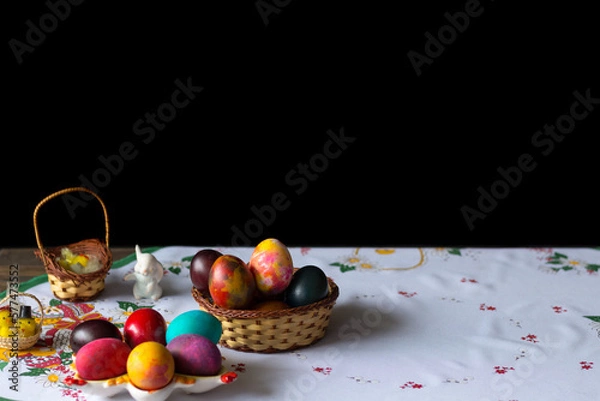 Obraz Colored easter eggs in a wooden basket at a white tablecloth on a black background
