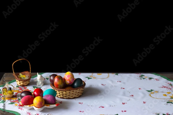 Obraz Colored easter eggs in a wooden basket at a white tablecloth on a black background