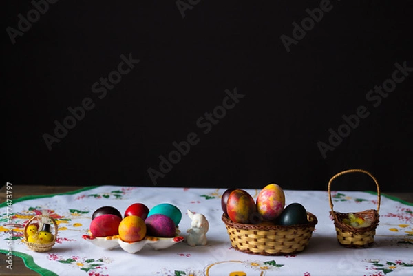 Obraz Colored easter eggs in a wooden basket at a white tablecloth on a black background