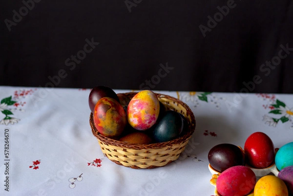Obraz Colored easter eggs in a wooden basket at a white tablecloth on a black background