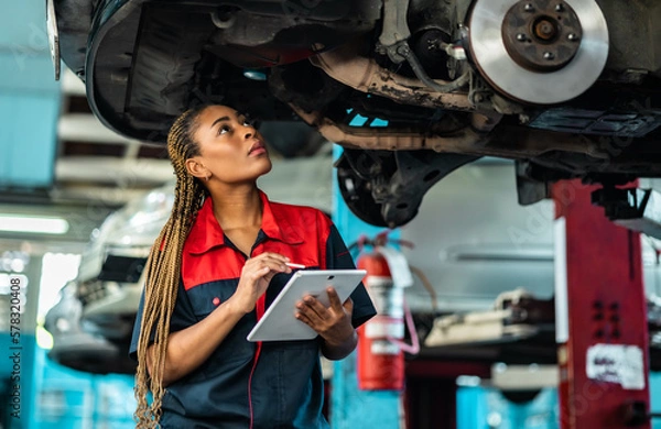 Fototapeta Engineer team checking under car condition on lifter in garage.Young auto mechanic in uniform is looking at camera and smiling examining car...
