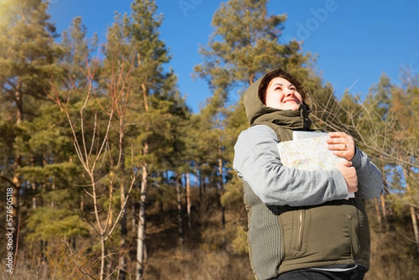 Obraz Photo from below of a traveler woman in winter clothes with a hood on her head looking at the sun and holding a map for trip in her hands.