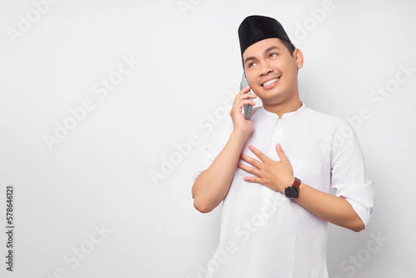 Obraz Friendly smiling young Asian Muslim man in Arabic clothes talking on mobile phone with hand his chest with grateful expression isolated on white background. People religious Islamic lifestyle concept