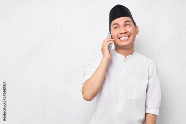 Obraz Portrait of smiling handsome young Asian Muslim man wearing Arabic clothes talking on mobile phone and looking empty space isolated on white background. People religious Islamic lifestyle concept
