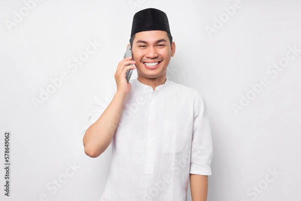Obraz Portrait of smiling handsome young Asian Muslim man wearing Arabic clothes talking on mobile phone and looking at camera isolated on white background. People religious Islamic lifestyle concept