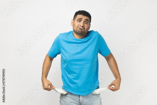 Fototapeta Sad poor attractive man in T- shirt standing showing his empty pockets, has no money, lost his job.
