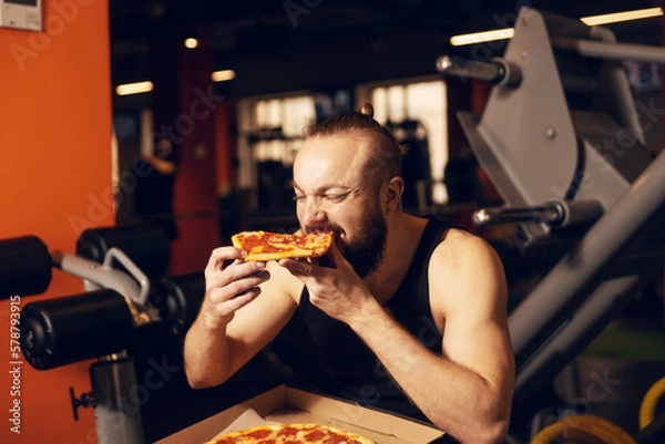 Obraz A young man with a beard is happy to eat pizza in the gym.