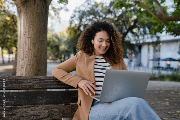 Obraz pretty curly woman sitting in city street in stylish jacket, working remote job on laptop