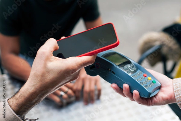Fototapeta Hand of a caucasian young man approaching a smart phone to a dataphone, close up. Paying bill using NFC technology. Concept of contacless payment