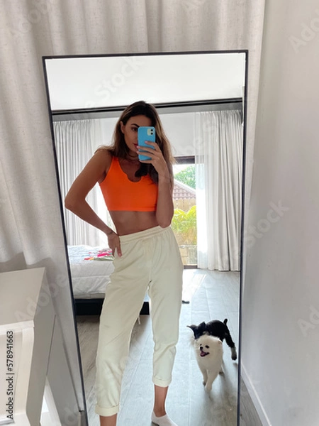 Fototapeta Fit tanned woman in orange top and cosy comfy pants at home take photo selfie on phone in mirror for social media, stories, vertical.