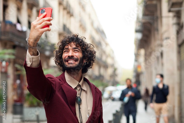 Fototapeta Handsome young man taking a self portrait while walking through the city. Urban millenial with beard and earring using a smart phone and smiling carefree outdoors. Copy space.