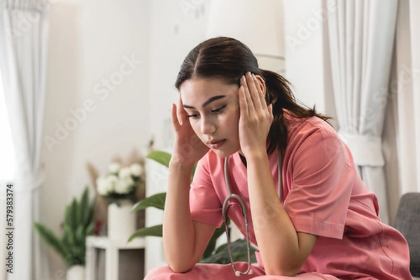 Obraz Upset female nurse sitting on a couch at home. Healthcare worker having headache. Doctors face heavy levels of stress. Young Caucasian caregiver in pink scrubs sitting hand touching temple tired face.