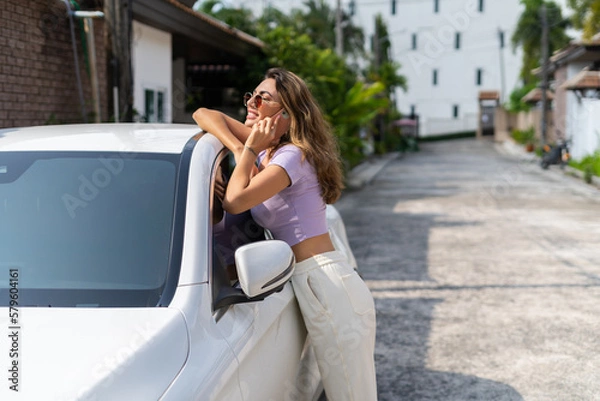 Fototapeta Successful smiling fit tanned attractive woman in casual wear is using her smart phone while standing near luxury modern car outdoors.