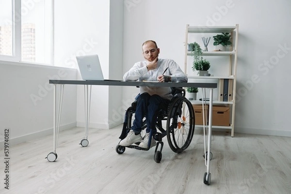 Fototapeta A man in a wheelchair, a businessman in the office works at a laptop, writing down a plan in a notebook, thoughtfulness, integration into society, the concept of working a person with disabilities